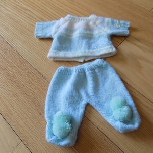 Könnte beinhalten: Ein gestricktes Baby-Outfit in Hellblau und Weiß. Das Oberteil hat kurze Ärmel und ein gestreiftes Design. Die Hose ist hellblau mit Bommeln an den Füßen. Das Outfit ist aus weichem Garn.