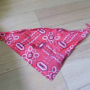 May include: A triangular bandana in a vibrant red colour featuring a repeating white and black geometric paisley-style pattern. The fabric is folded into a point, displaying a classic bandana print with intricate borders and central motifs.