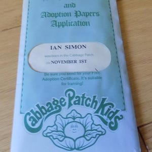 Peut inclure: Une enveloppe bleu clair pour un certificat de naissance et une demande de papiers d'adoption Cabbage Patch Kids. Le texte indique Ian Simon was born in the Cabbage Patch on November 1st. Elle comporte un dessin de chou avec un visage.