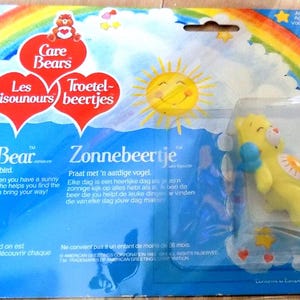 May include: A yellow Care Bear plush toy named Funshine Bear with a bluebird friend. The Care Bear is wearing a yellow and orange outfit with a heart-shaped sun on its chest. The packaging features a rainbow, clouds, and hearts. The text on the packaging reads "Care Bears", "Les Bisounours", "Troetel-beertjes", "Funshine Bear", "Zonnebeertje", and "Grosjojo".