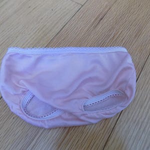May include: Light pink underwear with white lace trim along the waistband and leg openings. The underwear is made of a soft, smooth fabric and is laying on a wooden surface.