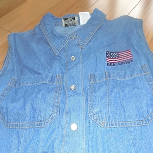 May include: A blue denim sleeveless shirt with a button-up front and two pockets. The shirt has a small American flag patch with the text "USA SPORT" below it.