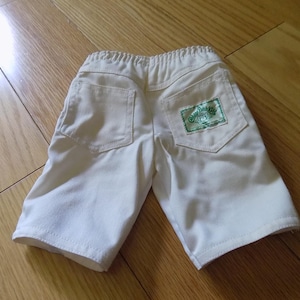 Może przedstawiać: Kremowe szorty z elastycznym pasem, dwiema tylnymi kieszeniami oraz prostokątną zielono-białą metką na prawej kieszeni z napisem Cabbage Patch Kids. Szorty posiadają widoczne przeszycia wzdłuż szwów oraz przy kieszeniach.