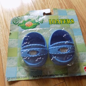 Puede incluir: Un par de zapatos de muñeca de plástico azul con una hebilla plateada. Los zapatos están empaquetados en una tarjeta con el logotipo de Cabbage Patch Kids y la palabra "Loafers". La tarjeta también dice "Shoes fit 35.6 cm (14") Cabbage Patch Kids Only!" y "For Ages Over 3 Years".