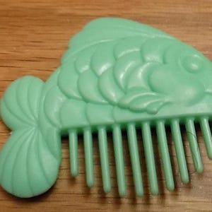 May include: A green plastic comb shaped like a fish. The comb has a detailed fish design and a row of teeth on the bottom.