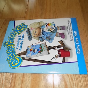 May include: A Cabbage Patch Kids coloring and activity book with a blue cover featuring a doll painting on an easel. The book title is "Rainy Day 'Kids".