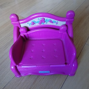 May include: A bright purple Fisher-Price toy bed with a tufted seat and a decorative headboard featuring a white panel with purple berry and leaf accents. The bed has rounded posts and a rectangular base.