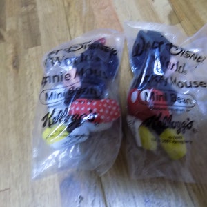 May include: Two plastic bags containing small plush toys of Mickey Mouse and Minnie Mouse. The bags are labeled "Walt Disney World" and "Mini Bean" and feature the Kellogg's logo.