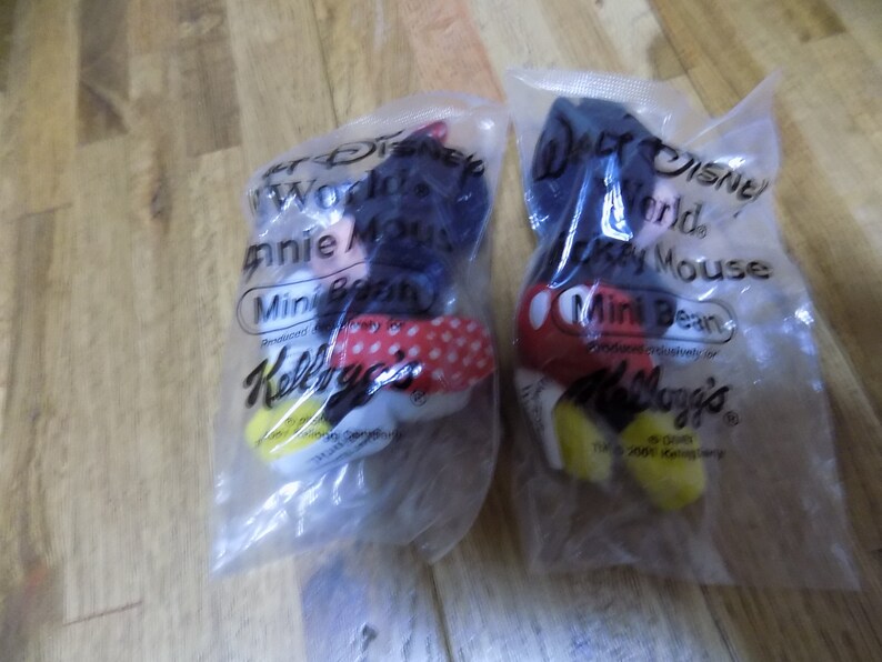 May include: Two plastic bags containing small plush toys of Mickey Mouse and Minnie Mouse. The bags are labeled "Walt Disney World" and "Mini Bean" and feature the Kellogg's logo.