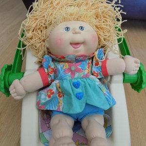Peut inclure: Une poupée Cabbage Patch Kid aux cheveux blonds portant une robe bleue à fleurs avec des boutons bleus. La poupée est assise dans une balançoire pour bébé verte et blanche.