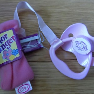 May include: Pink baby doll pacifier with a white flower design and a pink and white fabric strap attached to a pink fabric pouch with the text "Color Changer" and a CPK logo.