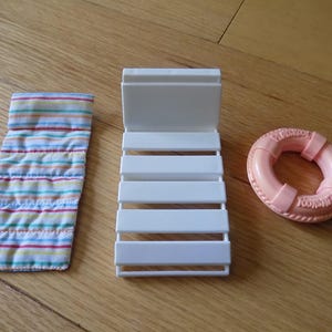 Puede incluir: Una tumbona en miniatura de plástico blanco, un cojín rectangular de tela con rayas horizontales en rosa, azul y amarillo, y un pequeño salvavidas redondo de plástico color melocotón con texto en relieve en su superficie.