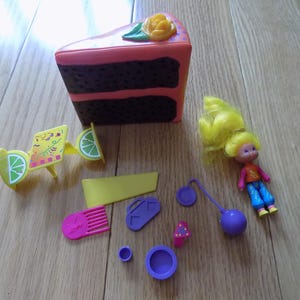 Puede incluir: Un juego de juguetes vintage con una porción de pastel, una muñeca pequeña con cabello amarillo y varios accesorios. El pastel es marrón y rosa con una rosa amarilla. Los accesorios incluyen una silla amarilla, un peine y otros pequeños artículos morados y rosas.