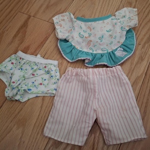 Könnte beinhalten: Ein Set aus drei Puppenkleidern, bestehend aus einer weißen und rosa gestreiften Shorts, einem weißen Unterhosen mit Blumendruck und einem türkisfarbenen und weißen Oberteil mit Rüschenbesatz.