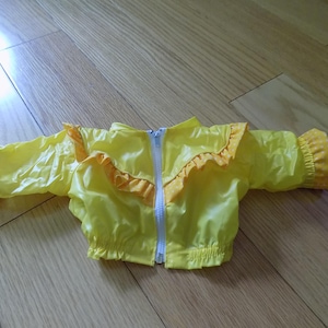 May include: A bright yellow jacket with a white front zip, featuring ruffled trim across the chest and sleeves with a white polka dot pattern on the cuffs. The garment has long sleeves and an elasticated hem, designed for a doll or small figure.