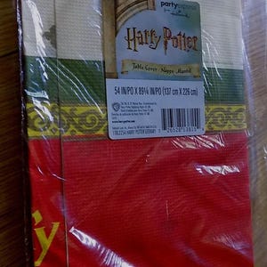 May include: A Harry Potter themed table cover in red, green, and gold with a decorative border. The table cover is 54 inches by 89 1/4 inches, or 137 centimeters by 226 centimeters.