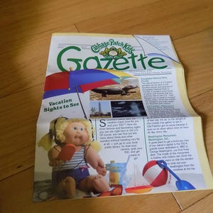 May include: A Cabbage Patch Kids Gazette newsletter featuring a headline about vacation sights. The page includes a graphic of a blonde doll with pigtails holding an orange popsicle under a multi-colored umbrella, alongside text and images of an alligator.