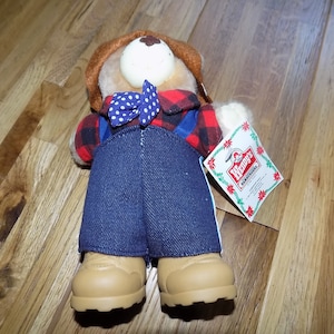 Puede incluir: Un oso de peluche marrón que lleva una camisa de cuadros rojos y negros, pantalones vaqueros azules, botas marrones y una pajarita azul y blanca de lunares. El oso sostiene una etiqueta que dice "Wendy's Hamburger".