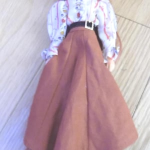 May include: A doll wearing a white blouse with a floral pattern, a brown skirt, and a black belt. The doll has long red hair and is wearing black shoes. The doll is standing on a wooden surface.