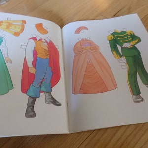 May include: A paper doll book with a red, green, and orange paper doll outfit. The paper doll is wearing a red cape, a yellow shirt, and blue pants. The paper doll is standing on a white background.