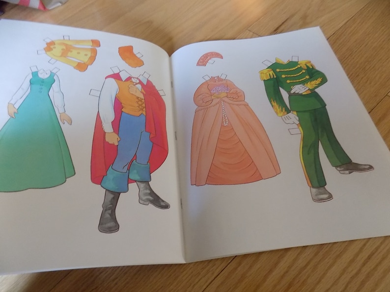 May include: A paper doll book with a red, green, and orange paper doll outfit. The paper doll is wearing a red cape, a yellow shirt, and blue pants. The paper doll is standing on a white background.