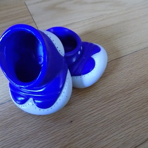 May include: A pair of blue and white plastic doll shoes. The shoes have a white trim and a blue base.