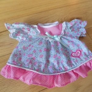 May include: A doll's dress with a floral pattern in light blue and pink. The dress features a pink polka dot ruffle, a heart-shaped patch with the letters "Cpk", and a light blue ribbon.
