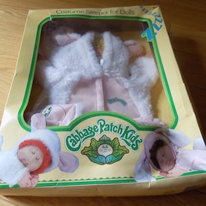 Op de afbeelding: Een Cabbage Patch Kids kostuum-slaappakje voor poppen in een gele kartonnen doos met een doorzichtig plastic venster. De verpakking bevat illustraties van poppen in dierenpakjes en de tekst Costume Sleeper for Dolls en Cabbage Patch Kids.