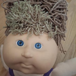 Könnte beinhalten: Eine Cabbage Patch Kid Puppe mit braunem und grünem Garnhaar, blauen Augen und einem lila und rosa Outfit.