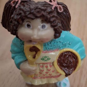 May include: A small figurine of a girl with brown hair in pigtails wearing a blue and white sweater, a yellow apron with pink hearts, and holding a yellow spatula and a brown chocolate chip cookie.