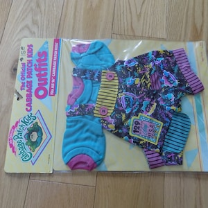 May include: A Cabbage Patch Kids outfit for a 16-inch doll. The outfit is a blue and pink shirt with a black and white patterned overall. The packaging includes the text "The Official Cabbage Patch Kids Outfits" and "Fits All 16" Cabbage Patch Kids".