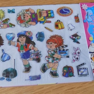 Può includere: Un set di adesivi Cabbage Patch Kids composto da un foglio di sticker colorati con vari personaggi, accessori come un boombox, uno zaino e un ciak cinematografico, e un'intestazione blu con il testo Cabbage Patch Kids cling set.