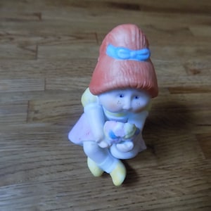 Puede incluir: Una pequeña figurita de cerámica de una niña con cabello rojo, que lleva un lazo azul y un vestido rosa. Ella sostiene un ramo de flores en sus manos.