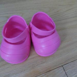 Puede incluir: Un par de zapatos de bebé de color rosa brillante con una correa en la parte superior. Los zapatos están hechos de un material liso y sólido y tienen una punta redondeada. Los zapatos están sobre una superficie de madera.