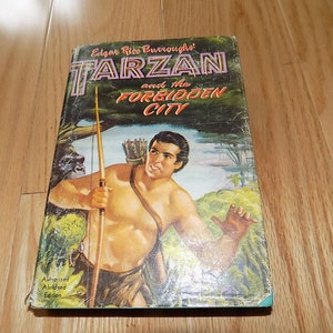 May include: A vintage paperback book cover featuring a muscular man holding a spear and bow and arrow. The cover is titled "Edgar Rice Burroughs' Tarzan and the Forbidden City" with an authorized abridged edition note.