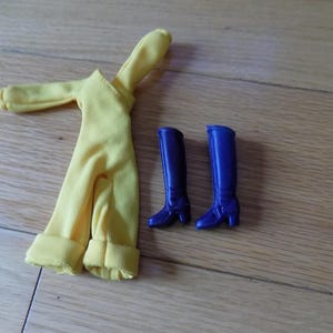 May include: A yellow doll jumpsuit with rolled cuffs and sleeves, paired with two pairs of blue knee-high boots. The boots have small heels. The items are arranged on a wooden surface.
