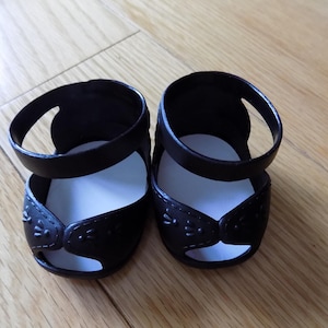 May include: A pair of black doll sandals with a strap around the ankle and a decorative design on the toe. The sandals have a white insole and are set on a wooden surface.