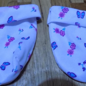 Może przedstawiać: Dwa białe buciki dla niemowląt z kwiatowym i motylkowym wzorem w odcieniach różu i błękitu. Buciki mają wywinięty mankiet u góry.