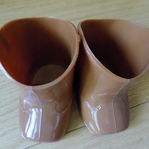 May include: A pair of small, brown, moulded plastic boots with a smooth, glossy finish. The boots feature a simple, rounded toe design and a wide opening at the top, showing a hollow interior. The construction includes visible seams along the sides.