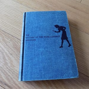 May include: A vintage book titled "The Mystery at the Moss-Covered Mansion" with a blue cover. The cover features a silhouette of a person holding a magnifying glass. The book is on a wooden surface.