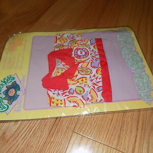 May include: A Cabbage Patch Kids fashion separates outfit for a doll. The outfit is a red and white shirt with a yellow, red, and blue print. The shirt has a red collar and a red button. The shirt is packaged in a clear plastic bag with a yellow and green background.