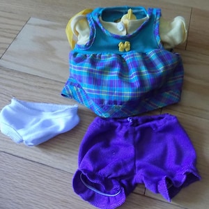 May include: A doll clothing set consisting of a sleeveless dress with a blue bodice, a yellow bow accent, and a purple and blue plaid skirt, a pair of solid purple shorts with elasticized leg openings, and a pair of plain white doll underwear.