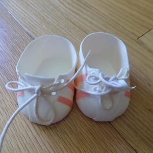 Puede incluir: Un par de zapatos pequeños de muñeca en color blanco con tres rayas finas de color coral en los laterales y cordones blancos atados en un lazo en la parte delantera. Los zapatos tienen una puntera redondeada y un acabado mate liso.