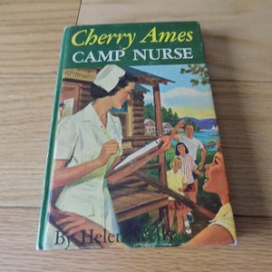 May include: A vintage book titled "Cherry Ames Camp Nurse" by Helen. The cover features an illustration of a nurse in a white uniform and cap, writing on a clipboard. A family is in the background near a wooden structure, with a lake and trees.