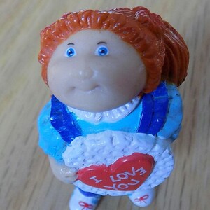 Könnte beinhalten: Eine rothaarige Cabbage Patch Kid Puppe, die ein blaues und weißes Kleid trägt und ein weißes herzförmiges Keks mit den Worten "I Love You" in Rot hält.