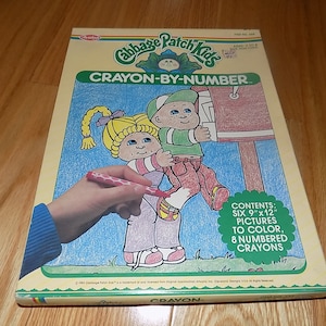 May include: A Cabbage Patch Kids Crayon-By-Number coloring book featuring six 9x12 inch pictures to color with 8 numbered crayons. The cover shows a hand holding a red crayon over a picture of two Cabbage Patch Kids.