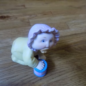 Puede incluir: Una pequeña figurita de cerámica de un bebé que lleva un mono amarillo y un gorro morado. El bebé está gateando y sosteniendo un juguete azul y rosa.