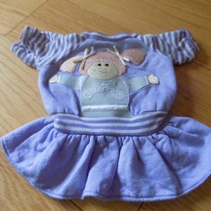 Könnte beinhalten: Ein hellviolettes Kleid mit kurzärmeligen weißen Streifen. Das Oberteil zeigt eine gestickte Cabbage Patch Kids-Figur mit rosa Haaren und weißen Schleifen. Die Taille hat ein weiß-violett gestreiftes Band über einem ausgestellten Rock.