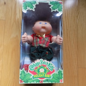 May include: A Cabbage Patch Kids Holiday Baby doll in its original packaging. The doll has brown hair, blue eyes, and wears a red and green plaid vest over a red shirt with dark green trousers. The box features holly leaf graphics and text.