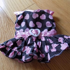 Puede incluir: Un vestido para perro negro y rosa con estampado de corazones. El vestido presenta una falda con volantes, un cinturón de satén rosa con una hebilla en forma de corazón blanca y un ribete de satén rosa alrededor del escote.